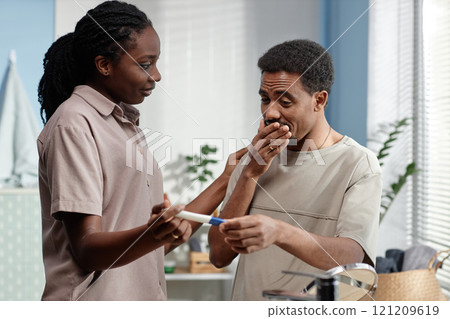 Medium shot of happy Black man holding pregnancy test looking in amazement at positive result surprised by wife at home 121209619