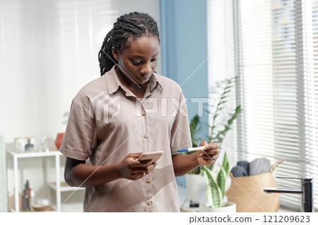 Serious young Black woman holding express pregnancy test and mobile phone waiting for result in bathroom, copy space Serious young Black woman holding express pregnancy test and mobile phone waiting for result in bathroom, copy space 121209623