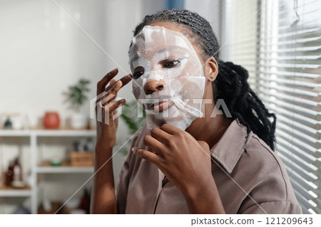 Young African American woman applying sheet mask saturated with nourishing ingredients on face lining it up with fingers while taking care of skin in morning at home, copy space Young African American woman applying sheet mask saturated with nourishing ingredients on face lining it up with fingers while taking care of skin in morning at home, copy space 121209643