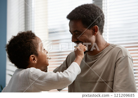 Happy Black man teaching little son dental hygiene while playful Black boy brushing fathers teeth in bathroom on Sunday morning 121209685