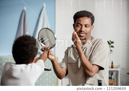 Medium shot of handsome Black man looking at reflection in mirror while little son helping dad during daily hygiene routine in bathroom Medium shot of handsome Black man looking at reflection in mirror while little son helping dad during daily hygiene routine in bathroom 121209689