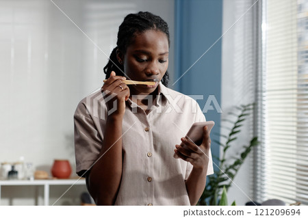 Medium shot of Black woman glued to phone screen while brushing teeth during self care routine in bathroom on Sunday morning, copy space Medium shot of Black woman glued to phone screen while brushing teeth during self care routine in bathroom on Sunday morning, copy space 121209694