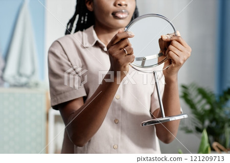 Cropped shot of young African American woman holding small mirror in hands applying makeup in morning at vanity table in bathroom, copy space 121209713