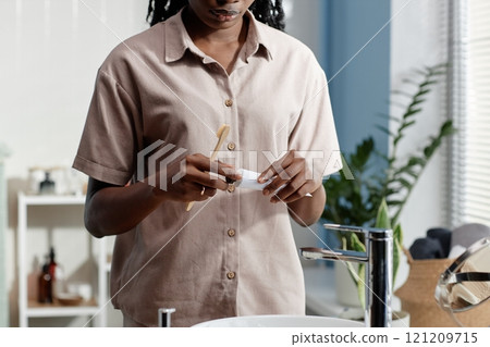 Cropped shot of young African American woman holding bamboo toothbrush and toothpaste tube taking care of dental health while standing at bathroom sink, copy space 121209715