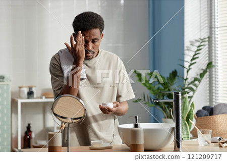 Medium shot of adult Black man applying cream on face taking care of skin while looking at reflection in mirror during morning routine in bathroom, copy space Medium shot of adult Black man applying cream on face taking care of skin while looking at reflection in mirror during morning routine in bathroom, copy space 121209717