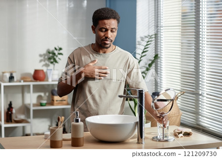 Medium shot of confident Black man with neat beard getting ready in bathroom while looking at reflection in mirror and admiring it in morning, copy space 121209730