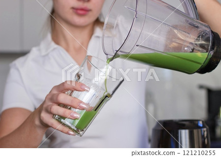 A girl in a white T-shirt pours a green smoothie into a glass. 121210255