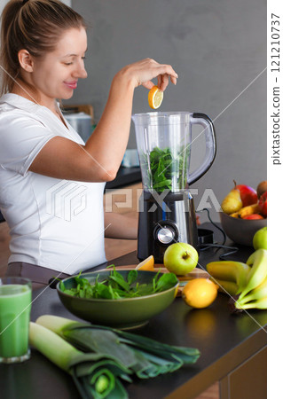 A girl in a white t-shirt throws spinach and lemon into a blender while standing in the kitchen A girl in a white t-shirt throws spinach and lemon into a blender while standing in the kitchen 121210737