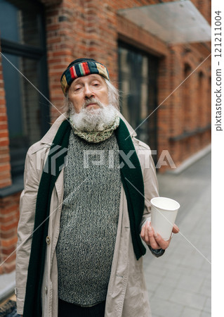 Vertical portrait of homeless senior man wearing worn-out coat, holding paper cup and begging for alms while standing on city street, looking at camera with sadness facial expression. 121211004