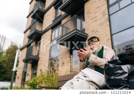 Low-angle portrait of aged homeless man sitting on city bench, using smartphone, trying to connect with family or access essential services. Concept of poverty and vulnerability, evoking empathy. Low-angle portrait of aged homeless man sitting on city bench, using smartphone, trying to connect with family or access essential services. Concept of poverty and vulnerability, evoking empathy. 121211014