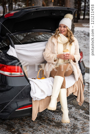 Young woman in woolen hat sits in trunk of car. Girl have a fun in the car. Winter travel, snow-covered trees in the forest. Snowy weather.  121211458