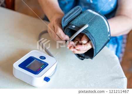 Senior woman finish to check blood pressure level at home, older female suffering from high blood pressure sitting and using a pulsometer, tonometer 121211467