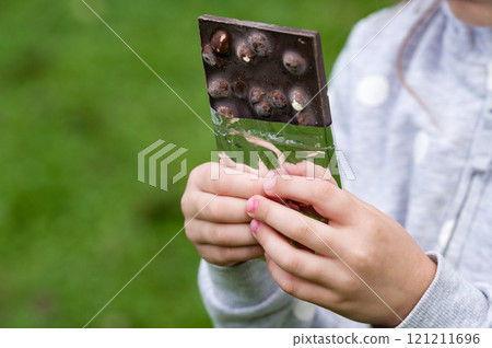 Child's hands holding a partially opened chocolate bar outdoors. 121211696