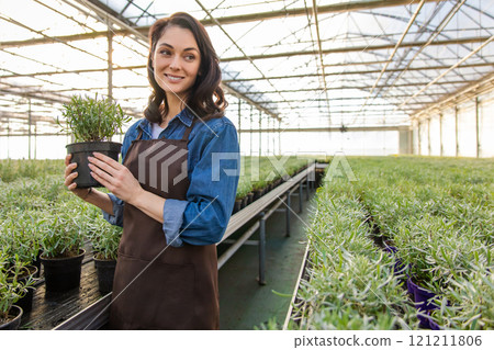 Dark-haired young woman standing in a greenhouse with a potted plant in hands Dark-haired young woman standing in a greenhouse with a potted plant in hands 121211806