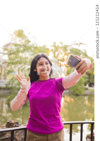 Young woman waving and smiling while making a video call with her smartphone in el retiro park 121212414