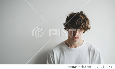 A young man with medium-length tousled hair wears a white shirt, looking down thoughtfully against a plain white backdrop, evoking a sense of calm A young man with medium-length tousled hair wears a white shirt, looking down thoughtfully against a plain white backdrop, evoking a sense of calm 121212964
