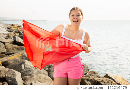 Young woman holding Morocco waving flag at coastline against of bright sea Young woman holding Morocco waving flag at coastline against of bright sea 121216479