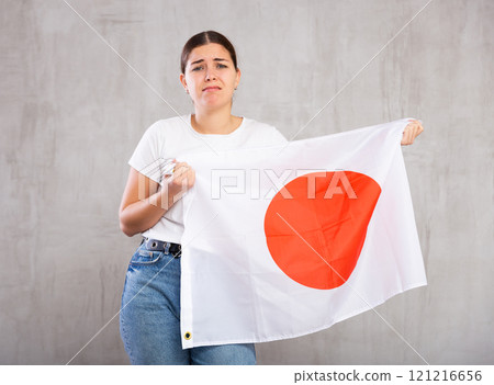 Disappointed young woman holding state flag of Japan against gray wall Disappointed young woman holding state flag of Japan against gray wall 121216656