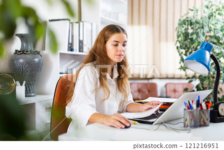 Focused young female office employee working with laptop and papers 121216951