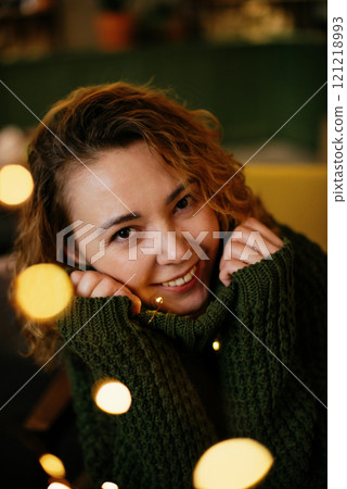 Cheerful middle-aged woman wearing a green knit sweater is surrounded by glowing Christmas and New Year garland lights, creating a cozy and festive holiday atmosphere 121218993