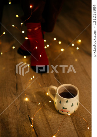 Female legs in red socks and mug of hot drink surrounded by glowing lights of Christmas and New Year, top view. Warm festive moment of woman surrounded by spirit of Christmas mood 121218994