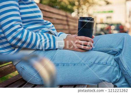 Woman relaxing on bench holding reusable coffee cup in park 121221262