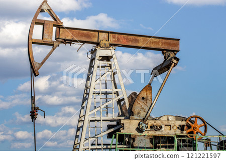 A weathered oil pumpjack, used for extracting oil, against a bright blue sky with scattered clouds. 121221597