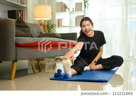 Smiling young woman doing hamstring stretch on a blue yoga mat in living room 121222668