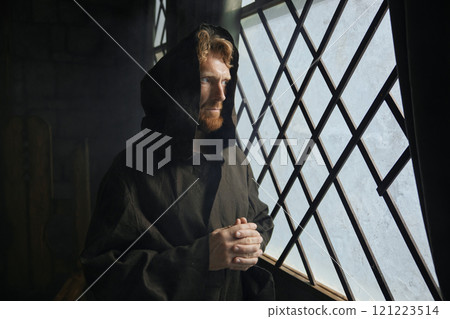Pensive monk in medieval costume with hood looking through window of monastery 121223514
