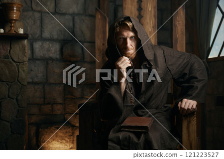 Portrait of medieval monk in hood sitting on wooden armchair 121223527
