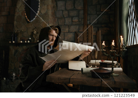 Medieval monk wearing robe with hood reading sacred manuscript on parchment 121223549