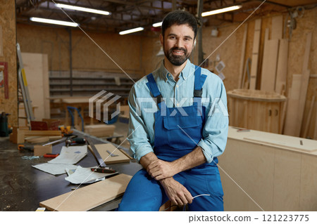 Skilled young adult woodworker sitting on workbench in well-equipped workshop 121223775