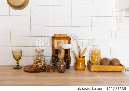 Kitchen counter with variety of items including wooden cutting board, bottle of wine, bottle opener, spoon, knife, bowl, jar, cup, vase and bowl 121225586