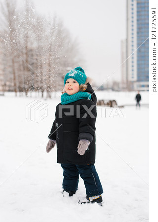 Boy child wearing a blue hat and a black coat is standing in the snow, throwing snowballs Boy child wearing a blue hat and a black coat is standing in the snow, throwing snowballs 121225591
