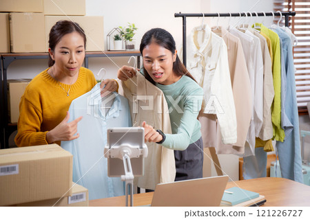 Two young asian woman live online clothing store while showing clothing with tablet on social. Two young asian woman live online clothing store while showing clothing with tablet on social. 121226727