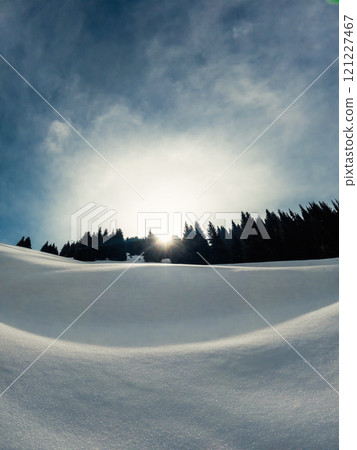 A wintry scene with snowcovered mountain slopes, trees in the background, and the sun peeking through the clouds, creating a picturesque natural landscape. 121227467
