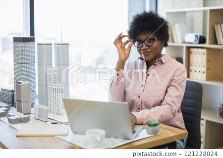 African woman architect in her 30s works on modern city model design with laptop. Office setting suggests creativity and professionalism in architecture field. African woman architect in her 30s works on modern city model design with laptop. Office setting suggests creativity and professionalism in architecture field. 121228024