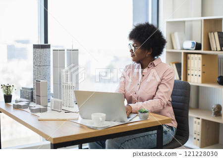 African woman architect designs modern city model. Middle-aged professional works on architectural project using laptop in bright office. Concept of creativity, innovation, urban planning. African woman architect designs modern city model. Middle-aged professional works on architectural project using laptop in bright office. Concept of creativity, innovation, urban planning. 121228030