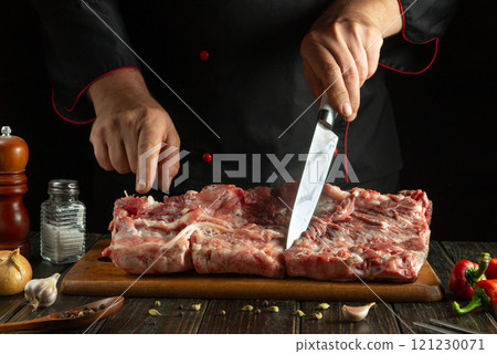 A skilled chef, dressed in a black apron, expertly slices fresh meat on a wooden board, surrounded by garlic, herbs, and spices. The atmosphere is warm and inviting, perfect for culinary creativity 121230071