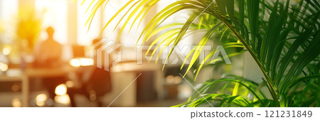 Blurred view of office space with employees working at desks on computers. Vibrant green palm leaves are in focus. Sunlight fills the room 121231849