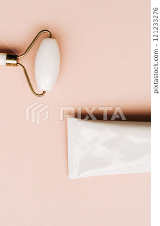 White jade facial roller and metallic cream tube with a black cap on a soft beige background 121233276