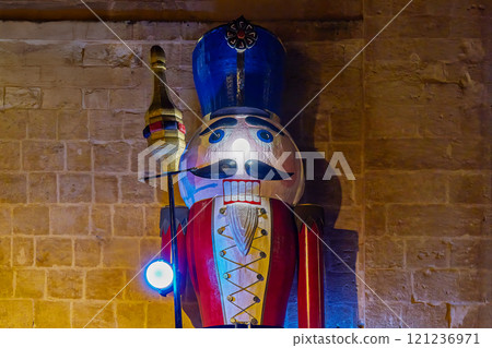 Wooden nutcracker soldier with bright red and yellow attire, illuminated by colorful blue and warm lights, stands against textured stone wall Wooden nutcracker soldier with bright red and yellow attire, illuminated by colorful blue and warm lights, stands against textured stone wall 121236971