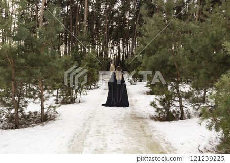 Blonde woman in a black cloak with a hood in winter in a pine forest. Female in cloak from the view from the back side Blonde woman in a black cloak with a hood in winter in a pine forest. Female in cloak from the view from the back side 121239225