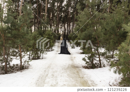 Blonde woman in a black cloak with a hood in winter in a pine forest. Female in cloak with hood from the view from the back side 121239226