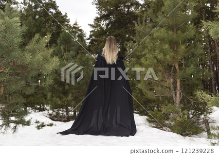 Blonde woman in a black cloak with a hood in winter in a pine forest. Female in cloak from the view from the back side Blonde woman in a black cloak with a hood in winter in a pine forest. Female in cloak from the view from the back side 121239228