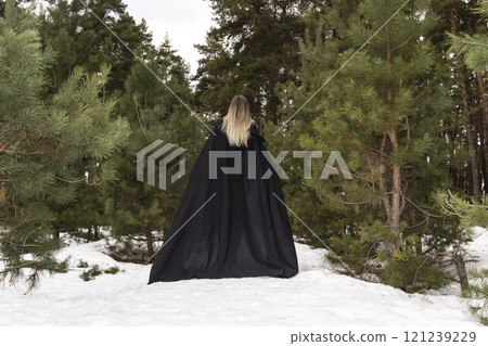Blonde woman in a black cloak with a hood in winter in a pine forest. Female in cloak from the view from the back side 121239229