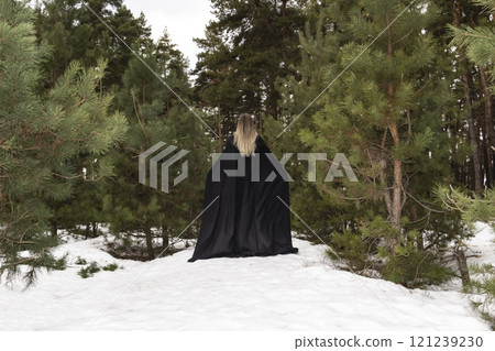 Blonde woman in a black cloak with a hood in winter in a pine forest. Female in cloak from the view from the back side Blonde woman in a black cloak with a hood in winter in a pine forest. Female in cloak from the view from the back side 121239230