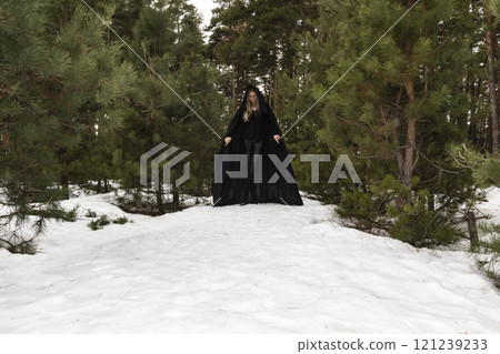 Blonde woman in a black cloak with a hood in winter in a pine forest 121239233