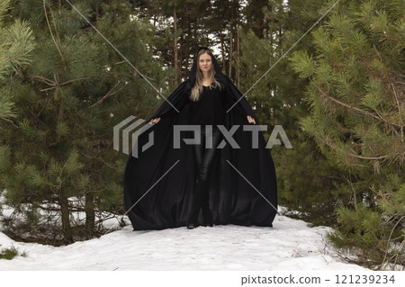 Blonde woman in a black cloak with a hood in winter in a pine forest Blonde woman in a black cloak with a hood in winter in a pine forest 121239234