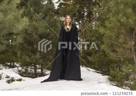 Blonde woman in a black cloak with a hood in winter in a pine forest Blonde woman in a black cloak with a hood in winter in a pine forest 121239235